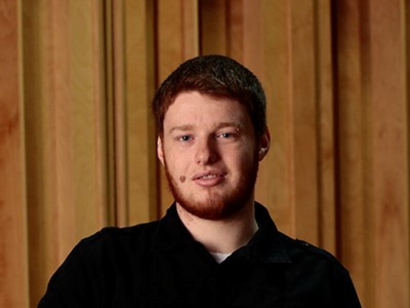 A young man with auburn hair and a beard, wearing a black shirt, stands in front of a wooden panel background.