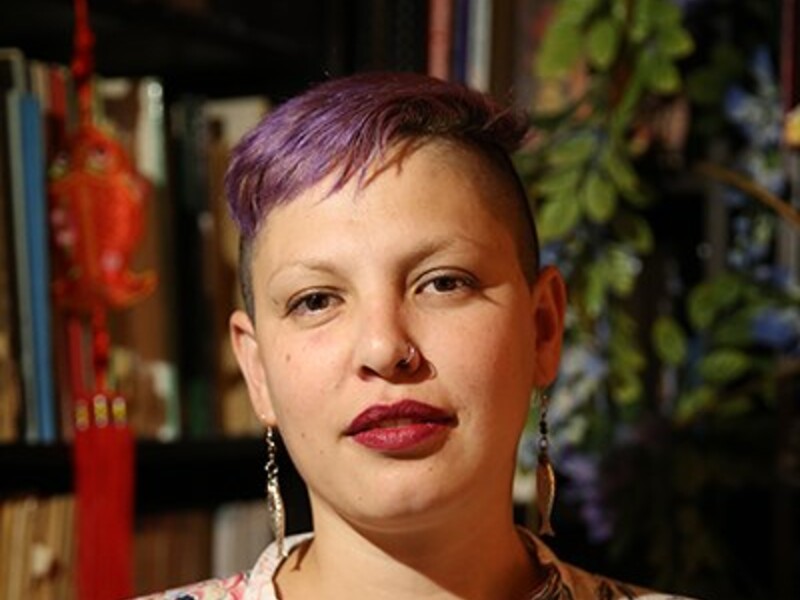 Person with short purple hair and floral shirt in a room with books and decorations in the background.