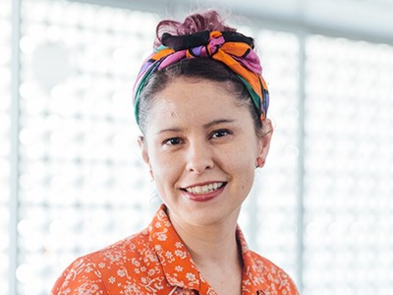 Person smiling indoors with a colorful headscarf and floral shirt.