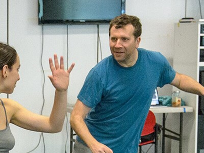 A man and woman engaged in a dance rehearsal in a studio, with a television and furniture in the background.