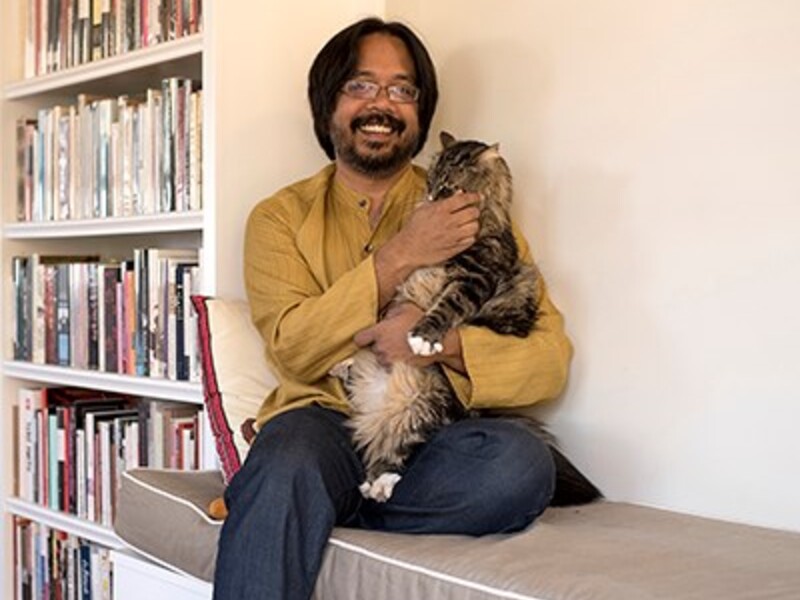 Person sitting on a cushioned bench holding a large fluffy cat, with a bookshelf in the background.