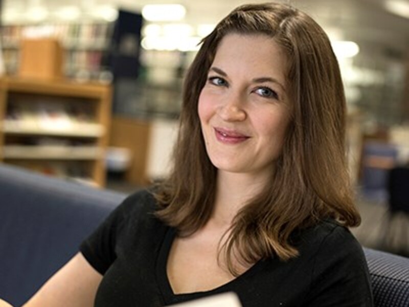 Person sitting in a library, holding a book and smiling at the camera.