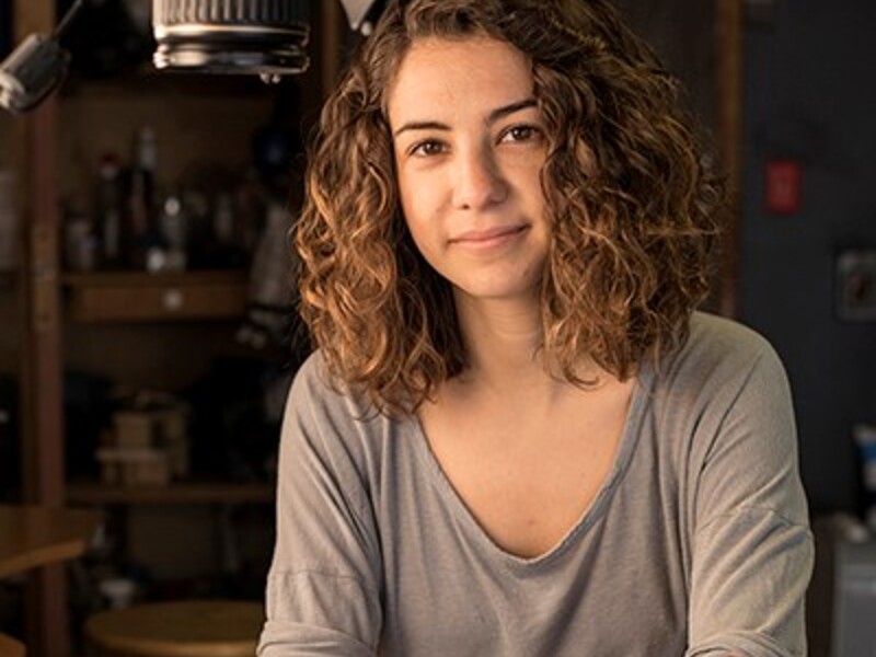 A person with wavy brown hair in a gray shirt sits at a table with photography equipment overhead.