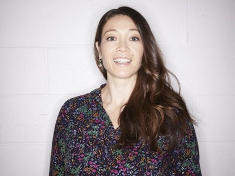 Person smiling in a floral blouse against a gray brick wall.