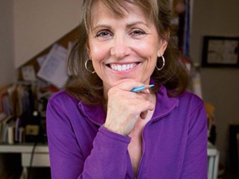 A person with light brown hair, wearing a purple top, smiles and holds a pen against their chin in a home office setting.