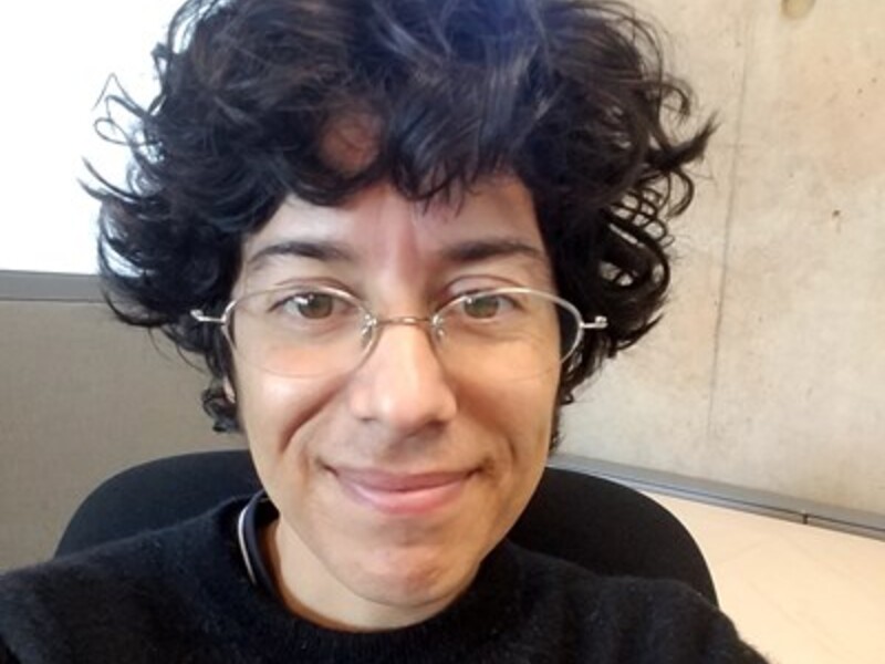 Person with curly hair and glasses smiling, sitting in front of a concrete wall and window.