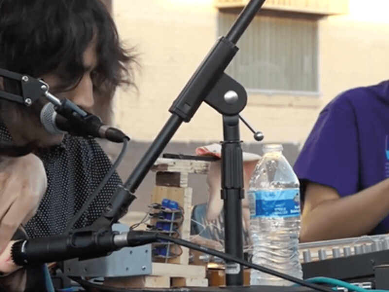 Two people seated at a table with electronic equipment and a microphone setup outdoors.