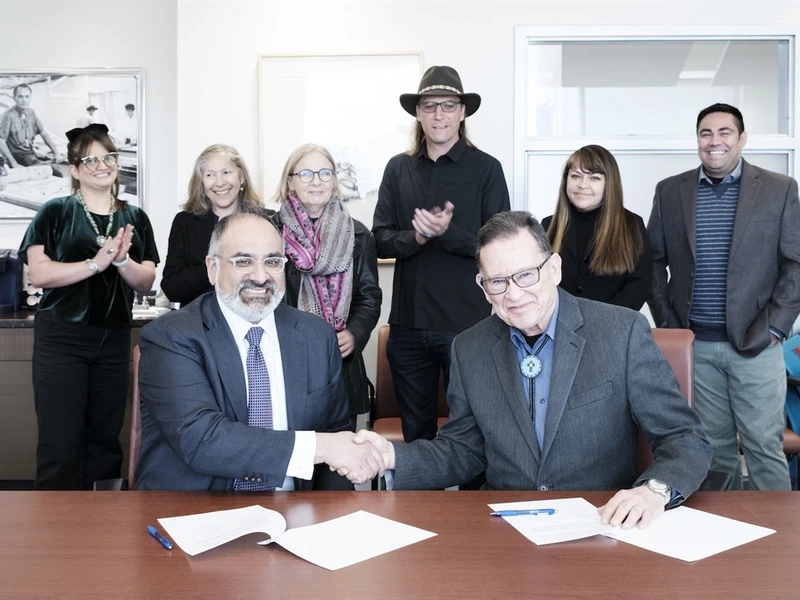 Two people in suits shaking hands at a table, surrounded by six smiling and clapping people.