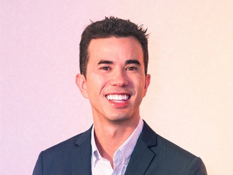 A man in a dark blazer and light shirt smiling against a warm gradient background.