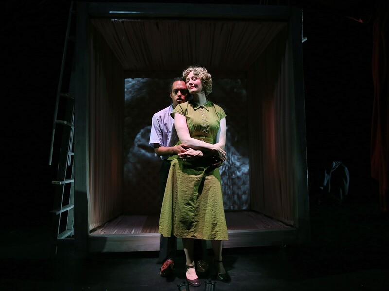 Two people stand closely together on a dimly lit stage, embraced within a box-like structure.