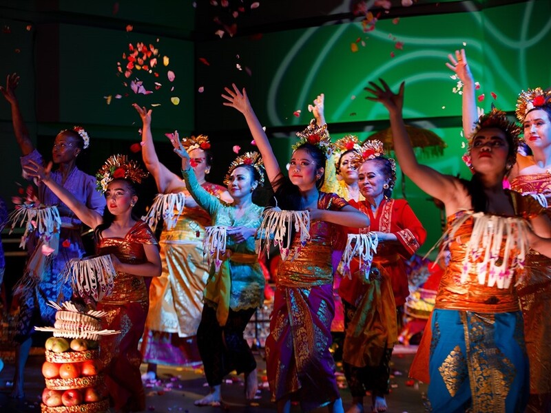 Dancers in colorful traditional Balinese costumes perform on stage, scattering flower petals.