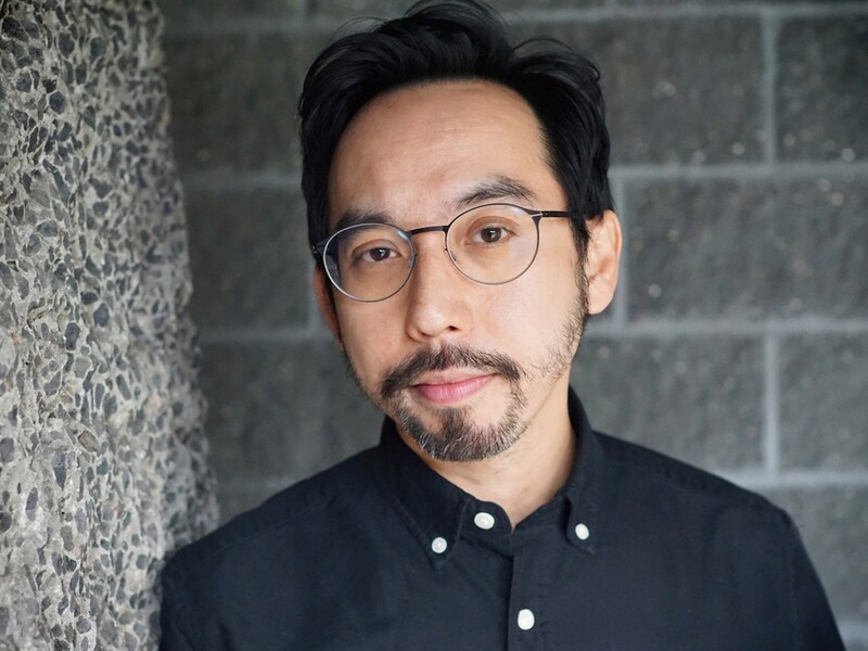 A person with glasses and a black shirt stands against a textured wall and brick background.