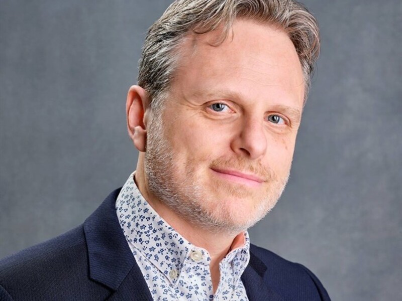 Man in a navy blazer with a white floral shirt against a gray background.