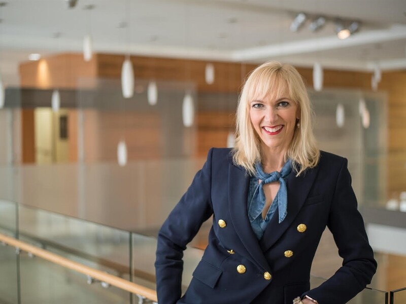 Smiling woman in a navy blazer with gold buttons in an office setting.