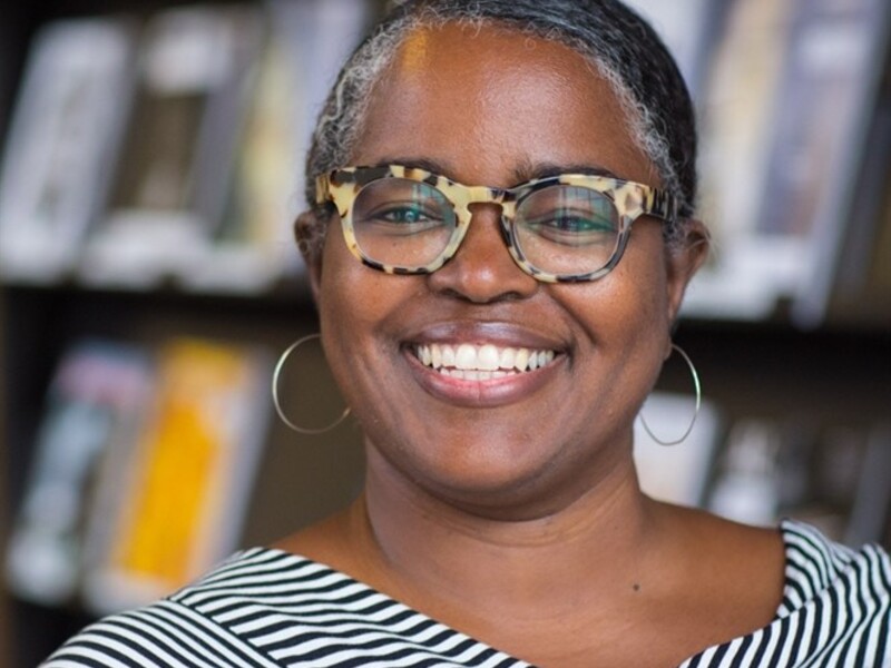 Smiling person with glasses and striped outfit in front of blurred shelves.