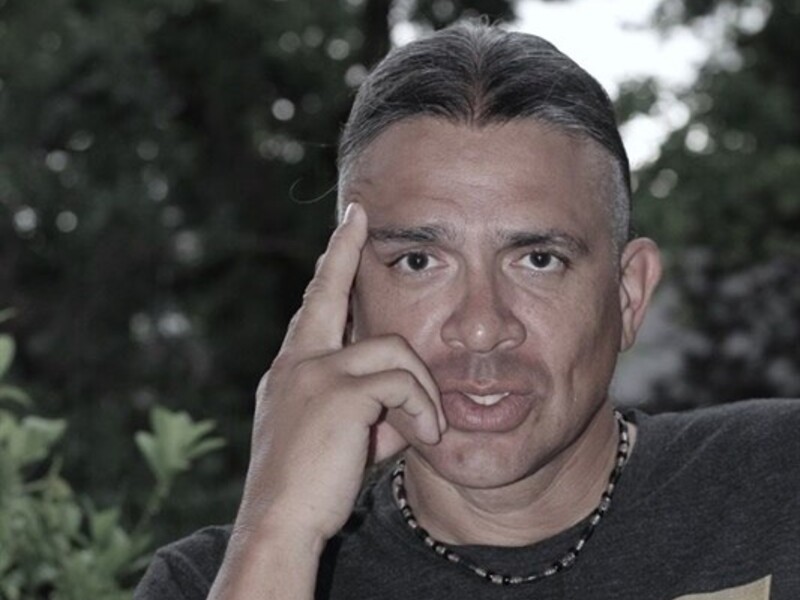 Person sitting outdoors with greenery in the background, wearing a dark t-shirt with a yellow "W" logo.