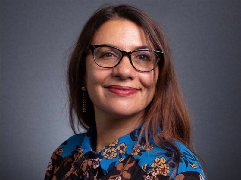 Portrait of a smiling person with glasses and a floral blouse against a grey background.