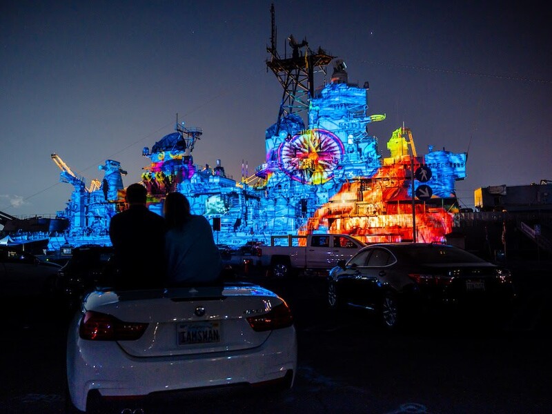 Large ship with colorful light projections at night, with silhouettes of two people sitting on a car in the foreground.