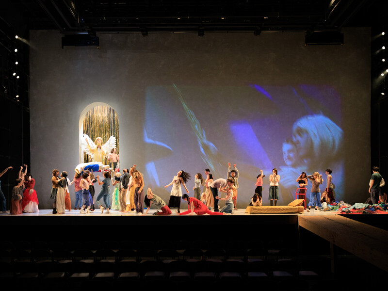 Actors performing on a stage with an angelic figure in a golden archway and a large projected image in the backdrop.