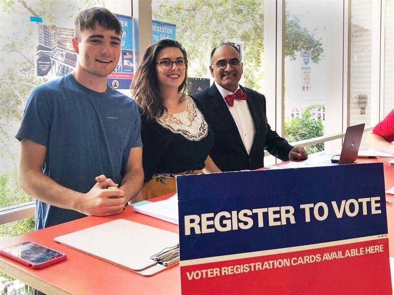 Three people standing behind a voter registration table with a "REGISTER TO VOTE" sign.