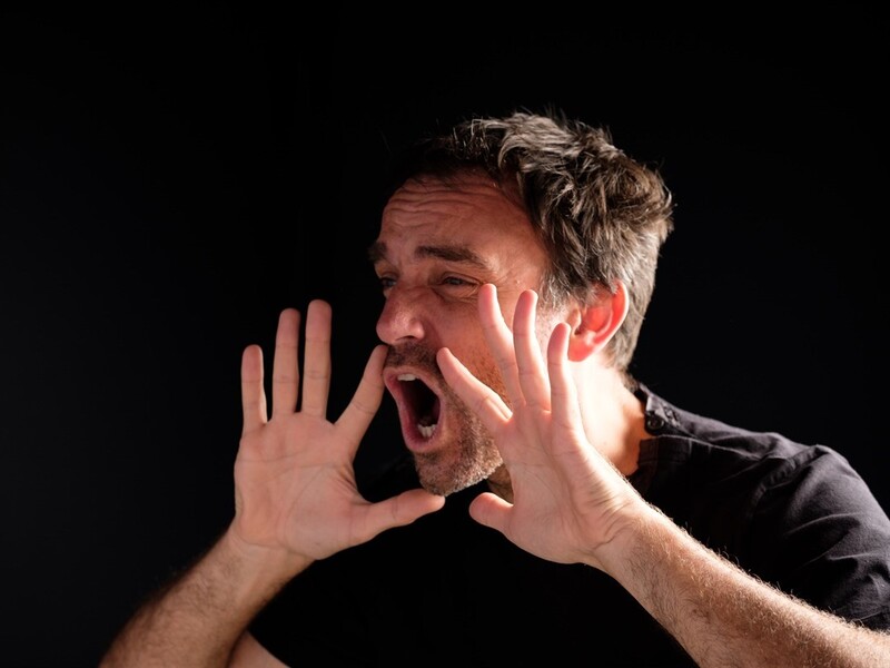 A person with raised hands and an open mouth, possibly shouting, against a dark background.