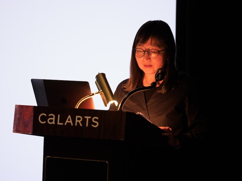 A woman reads at a podium in front of a white screen