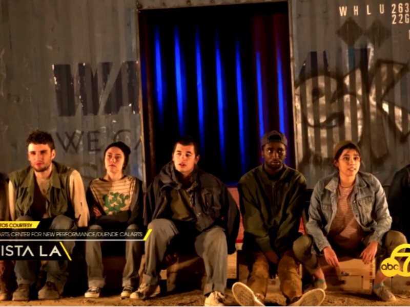 Seven people sitting on crates in front of a blue shipping container with graffiti.