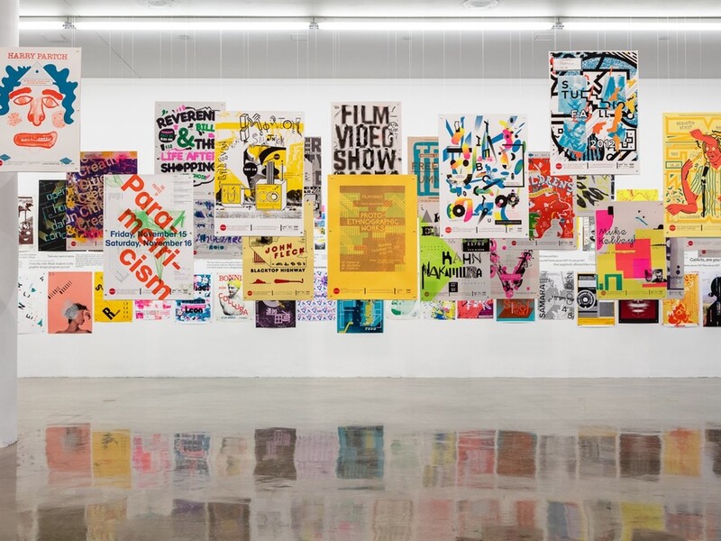 An art installation featuring a collection of colorful posters displayed on a white wall, each showcasing different designs and colors.