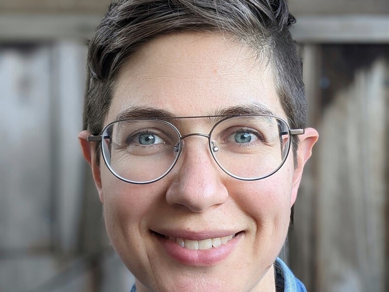 Close-up portrait of a person wearing glasses and a denim shirt, with a wooden background.