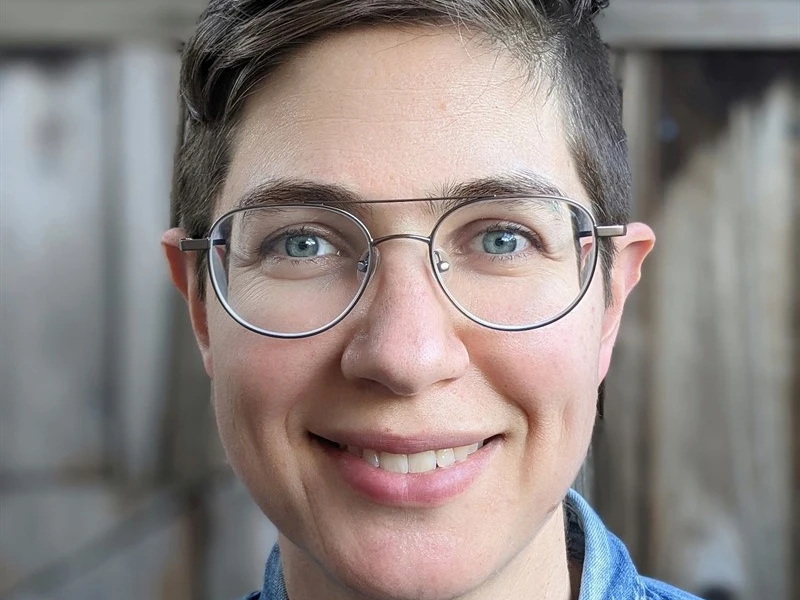 Close-up portrait of a person wearing glasses and a denim shirt, with a wooden background.