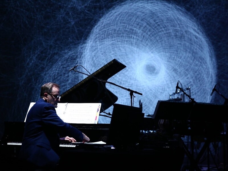 Person playing a grand piano in front of a swirling abstract projection.