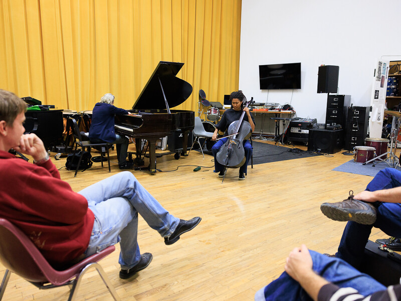 A student plays a cello in the center of a large practice room. Other students look on. 