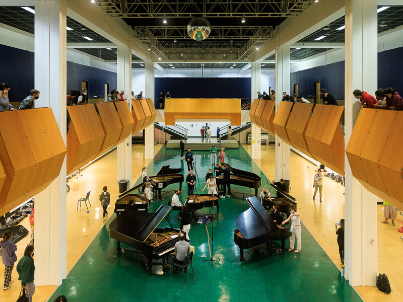 A circle of seven pianos in the main gallery at CalArts. Students look on from the balcony. 