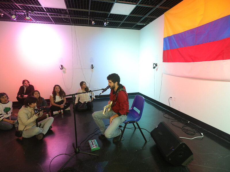 A person sits with a guitar and a microphone in a small room while a small group of people watch