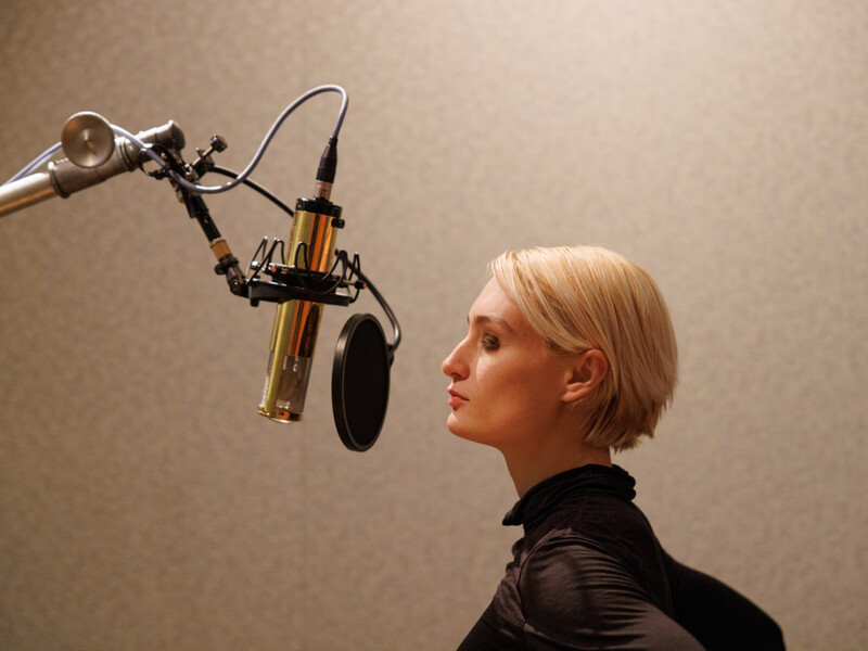 A singer with short blonde hair sings into a recording microphone