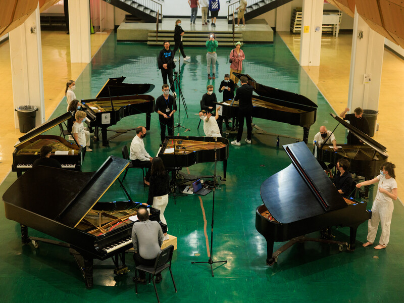 Seven pianos in a circle, each with players and dancers moving between them. 