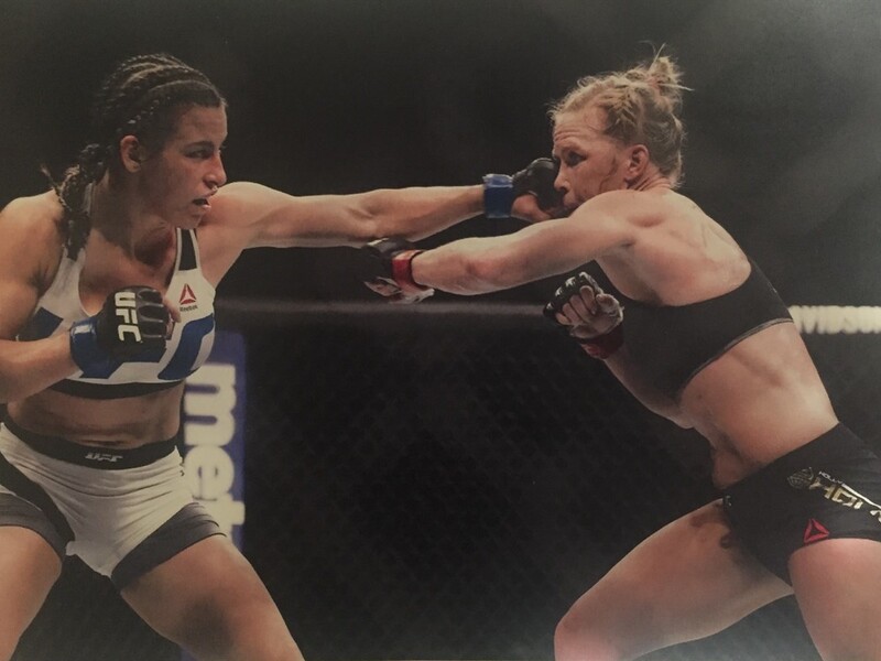 Two female mixed martial artists in a cage fight, one throwing a punch and the other blocking.