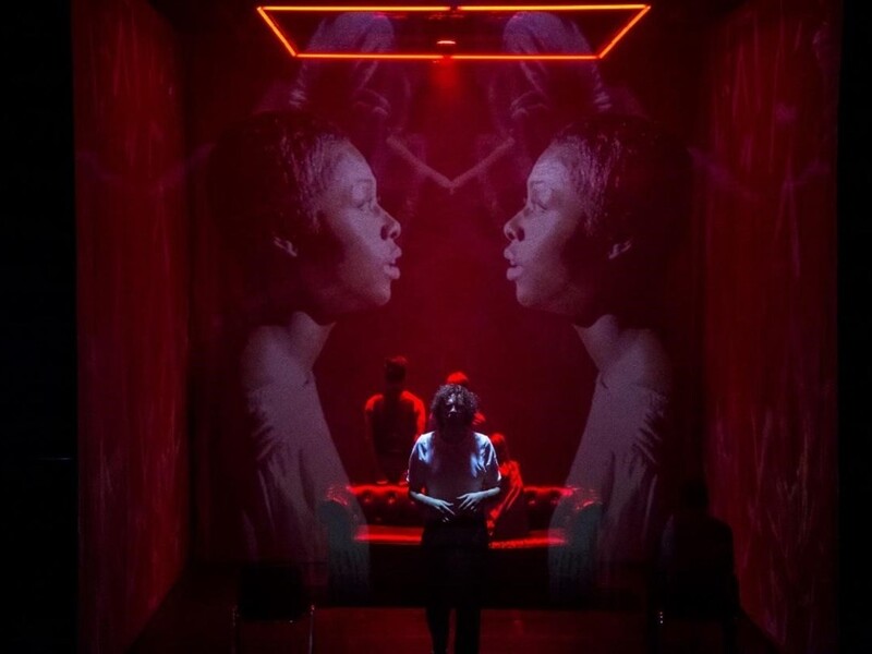 A dramatic red-lit scene with two large projected profile images facing each other and a person standing in front of a red couch.