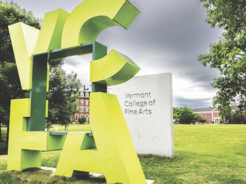 Abstract green sculpture spelling "VCFA" near Vermont College of Fine Arts sign with buildings and trees in the background.