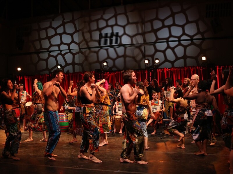 A group of performers dancing energetically on stage in colorful, patterned costumes, with musicians playing drums in the background.