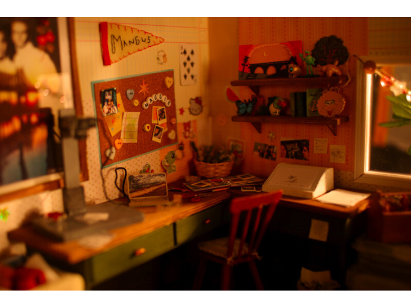 A cozy workspace with a cluttered desk, corkboard with pinned items, shelves with decorations, and warm lighting.