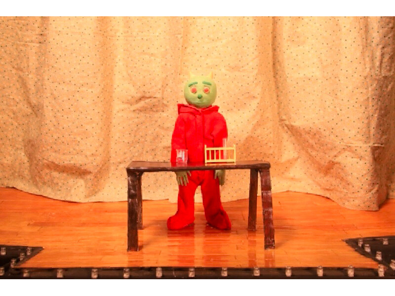 A puppet with a green face and red clothing stands behind a table on a stage with a polka-dot curtain backdrop.