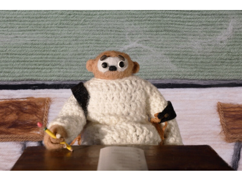 Felt puppet sitting at a desk in a classroom setting, holding a pencil.