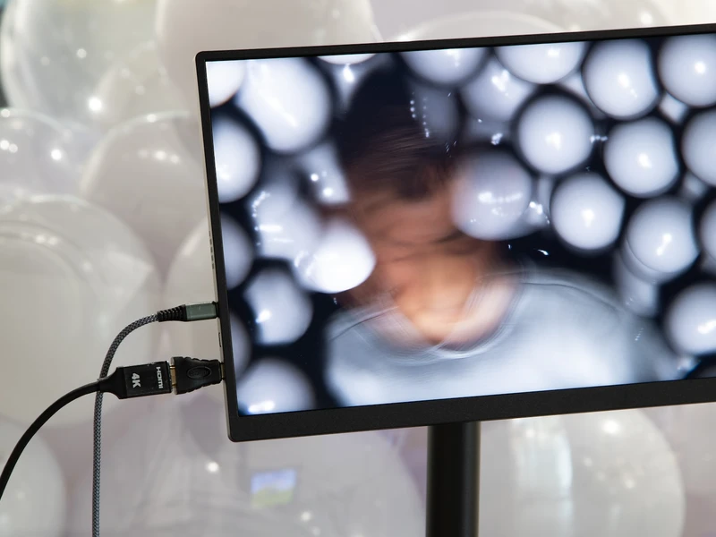 A monitor displays a blurred head amid a field of white balloons. Many white balloons are grouped behind the monitor.