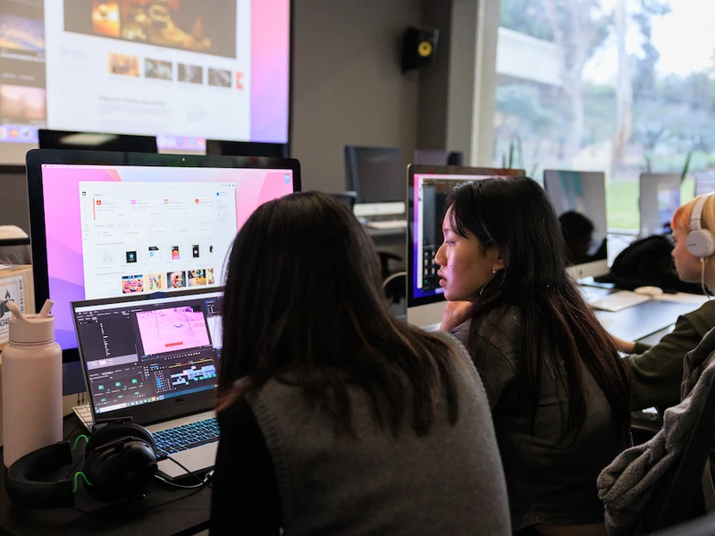 Two CalArts students huddle in front of a computer monitor, discussing their designs. 