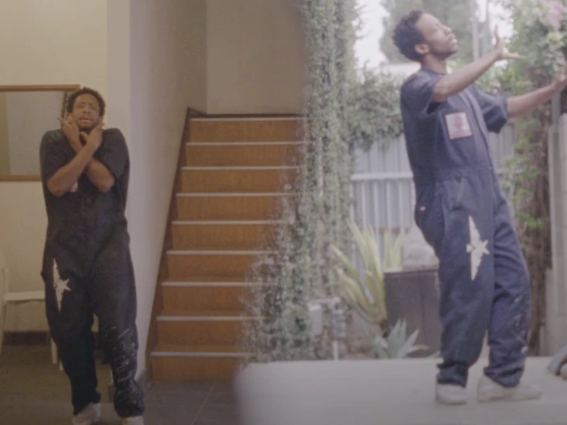 Person depicted indoors near a staircase on the left and outdoors beside a fence on the right, wearing a dark jumpsuit.