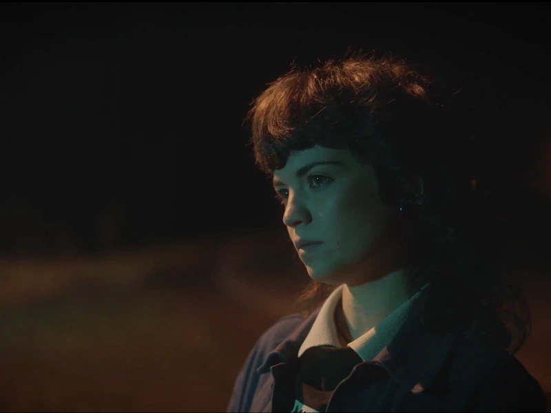 Person in profile with short dark hair, lit by bluish light against a dark background with an orange light in the distance.