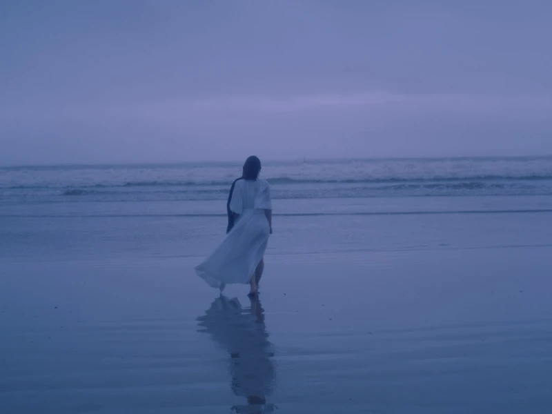 A person in a white dress stands on a beach facing the ocean under a blue, overcast sky.