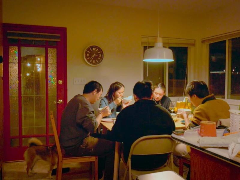 Dining room scene with six people eating at a wooden table, a dog near a red-framed door, and a clock showing 5:11.