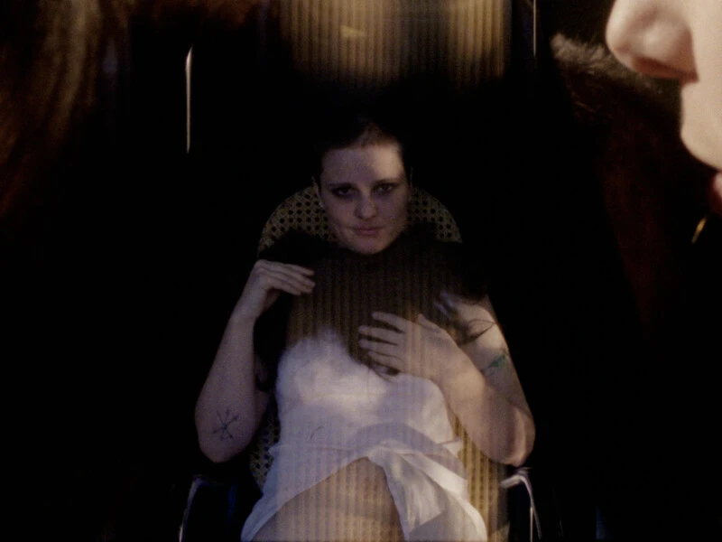 A person seated in a chair with a patterned back, in dim lighting, holding their hand to their chest.